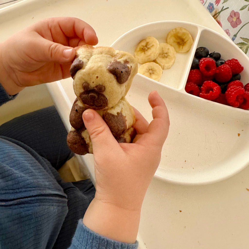 Kisgyermek két kézzel tart egy maci formájú sütit, háttérben gyümölcsös tányér