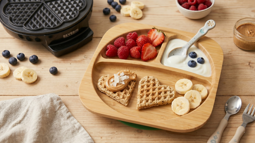 Baba gofri tálalva osztott bambusz tányéron – szív alakú gofri mogyoróvajas, bogyós gyümölcs és joghurt mellé