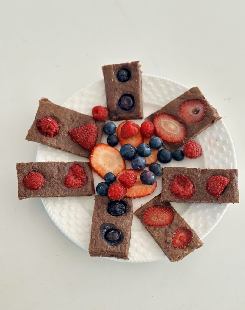 Cukormentes bababarát brownie szeletek fehér tányéron sugarasan elrendezve, közepén friss eper, málna és áfonya