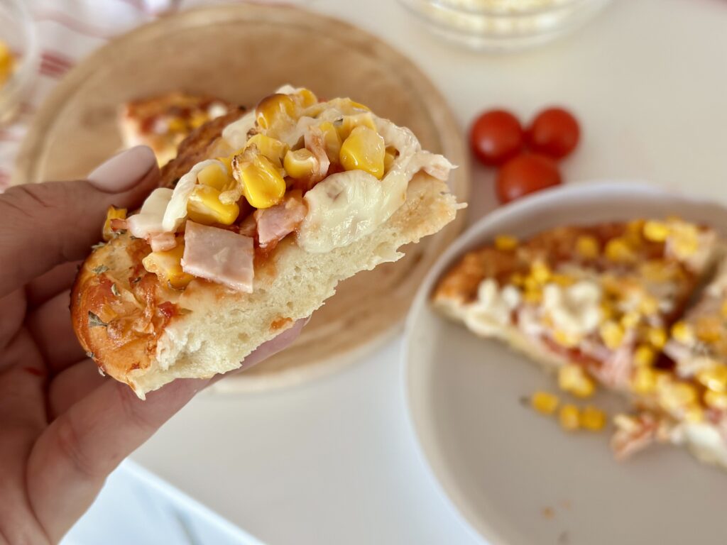 Kézben tartott pizzaszelet, puha levegős tésztával és sonkás-kukoricás feltéttel